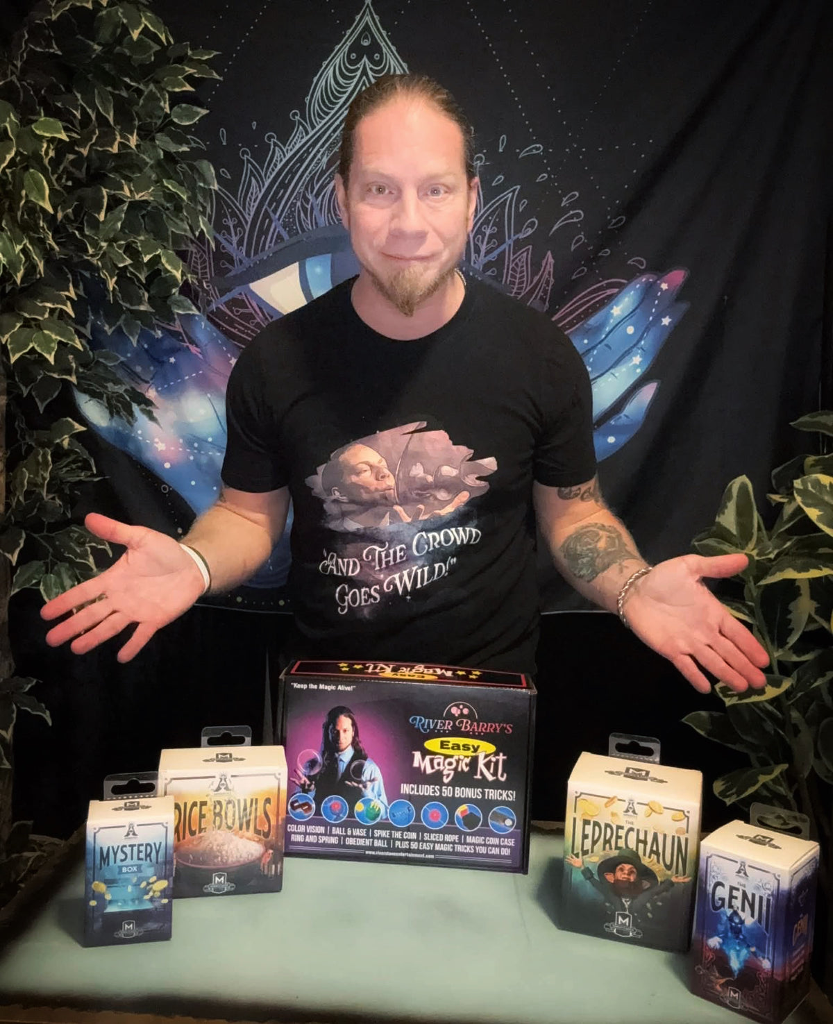 River Barry,, Man in a black t-shirt with a graphic design, standing behind a table with various boxes on a dark background. Magic kits and products.